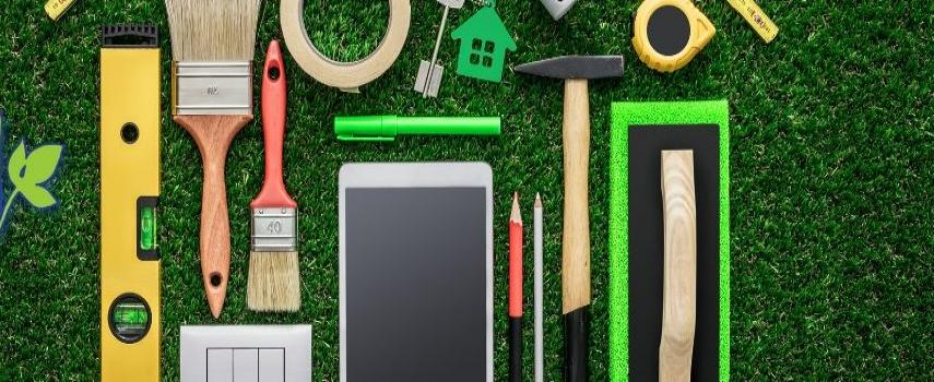 A flat lay of hardware tools, pencils, paintbrushes, an iPad, and tape on grass
