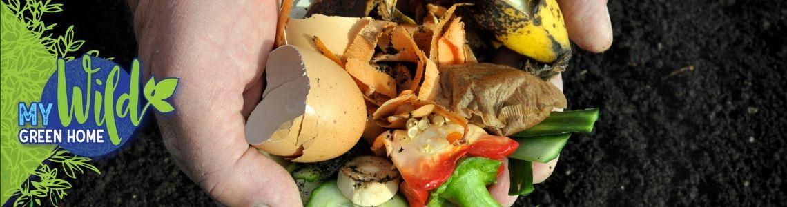 A handful of organic waste, including egg shells, banana peels, and vegetable scraps are held over a pile of dark brown dirt.